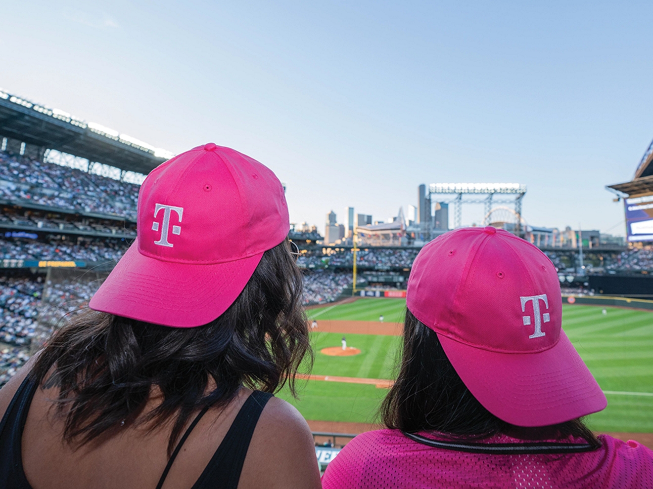 ​​Dos amigos ven un partido de béisbol con gorras magenta de T-Mobile.​